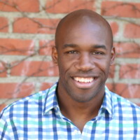 Nnamdi smiling to camera, wearing a blue and white checked shirt. He is standing in front of a red brick wall.